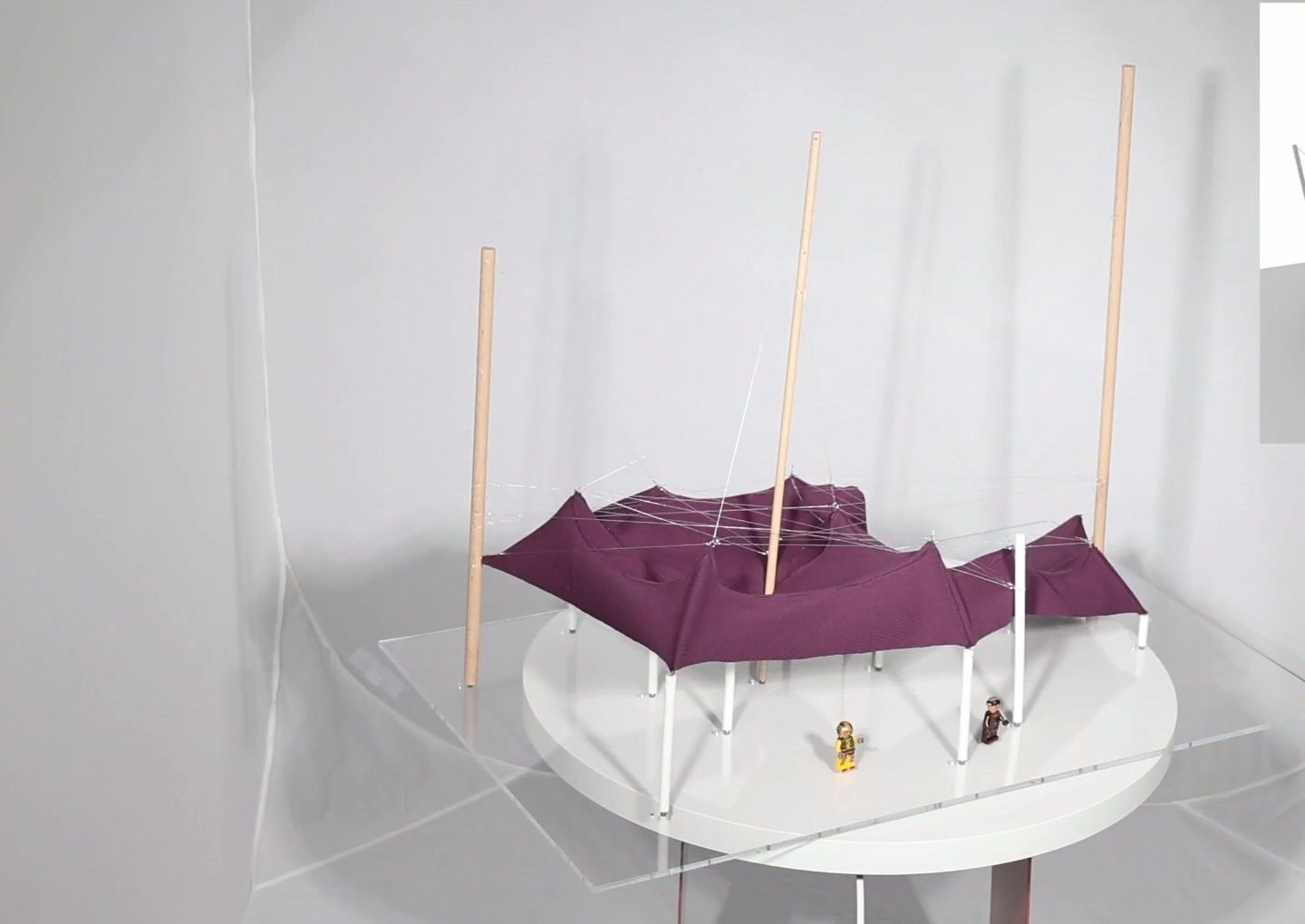 Physical prototype: a plum-coloured fabric tensile roof held by timber masts and cables, with tiny figures for scale